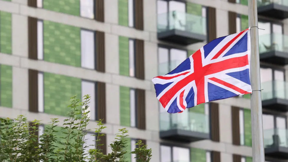 Union Jack flag waving in front of a modern building with greenery.
