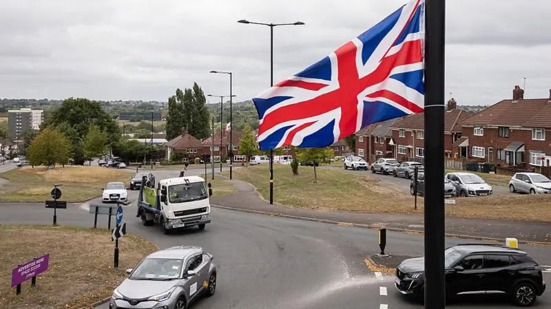 Street scene with a British flag, cars, and buildings on a cloudy day.